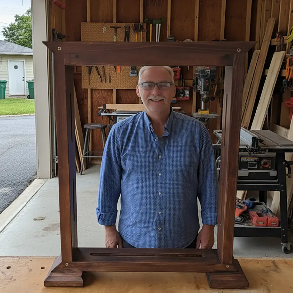 The owner and craftsman of StandCraft Canada smiling from behind a completed dark wood display stand in his Prescott, Ontario workshop